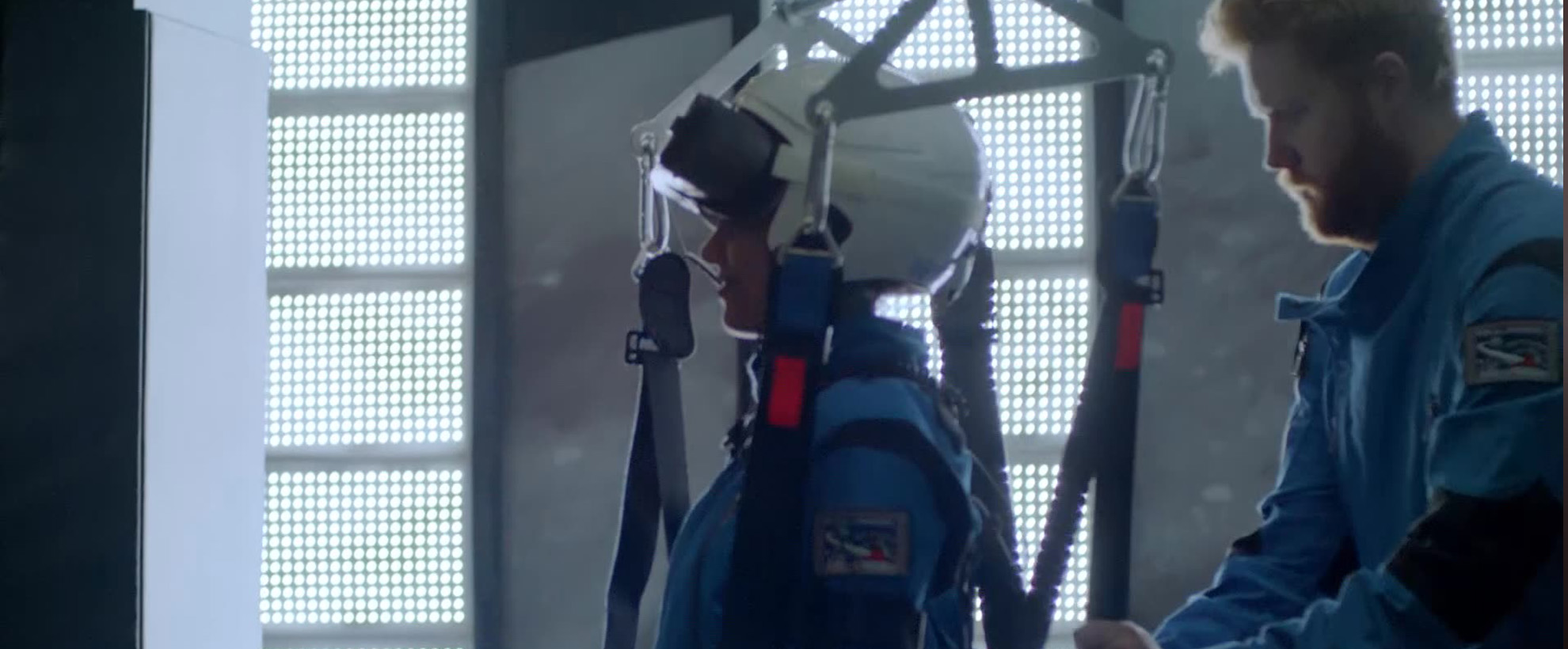 A woman in a white helmet and blue jumpsuit gets strapped into a harness by a red-headed man with a beard who is also in a blue jumpsuit