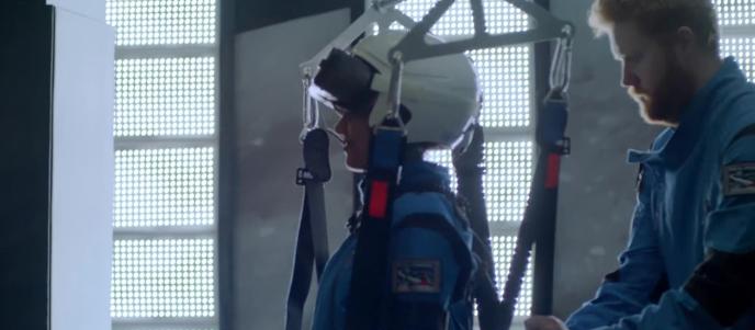 A woman in a white helmet and blue jumpsuit gets strapped into a harness by a red-headed man with a beard who is also in a blue jumpsuit