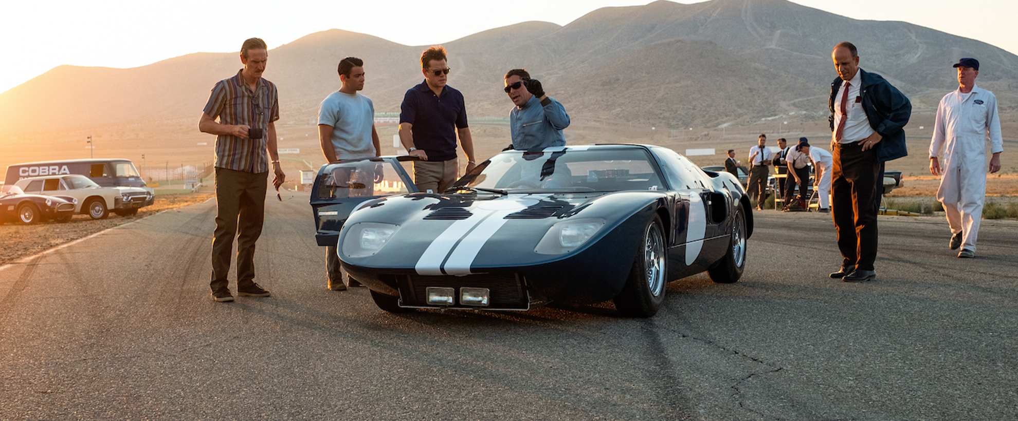 The cast of Ford vs Ferrari gather around a parked race car while the sun sets