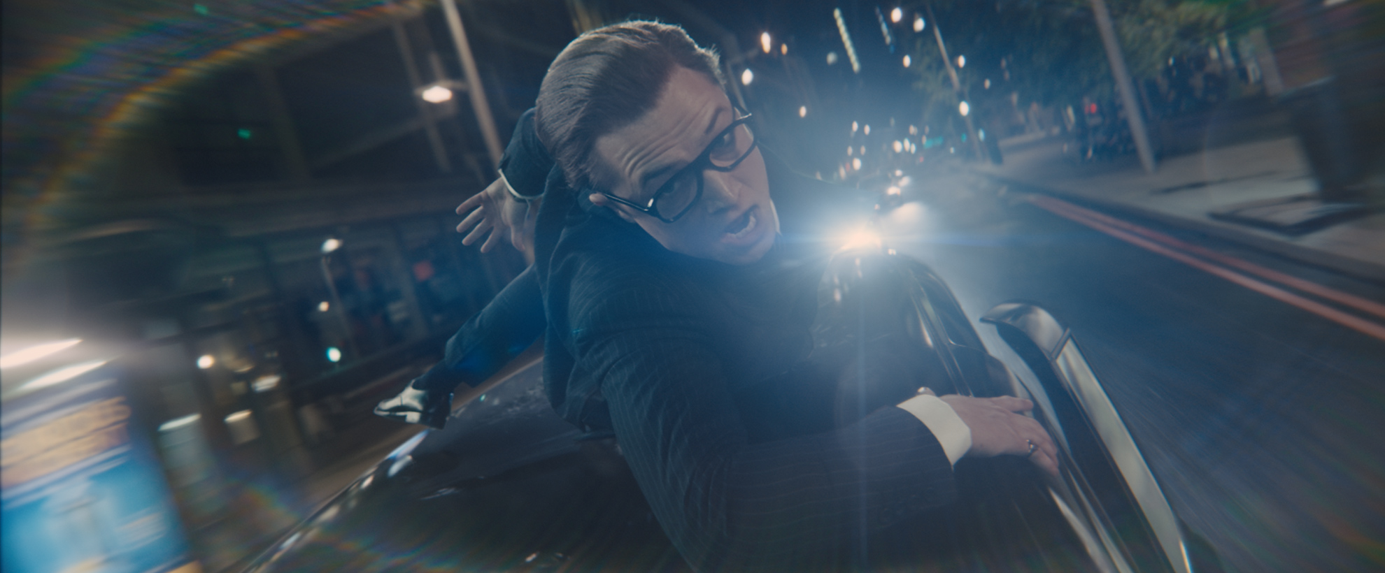 Eggsy (Taron Egerton) hangs on to the roof of a black taxi, speeding through London at night time