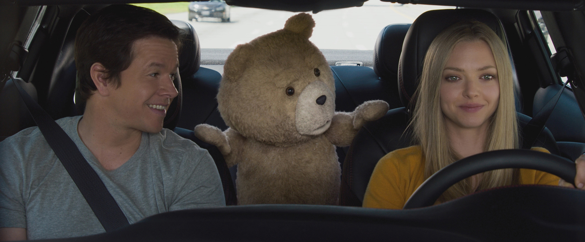 Amanda Seyfried drives a car with Mark Whalberg in the passenger seat. Ted, an alive teddy bear, pokes his head from the back seat to talk to the two in front