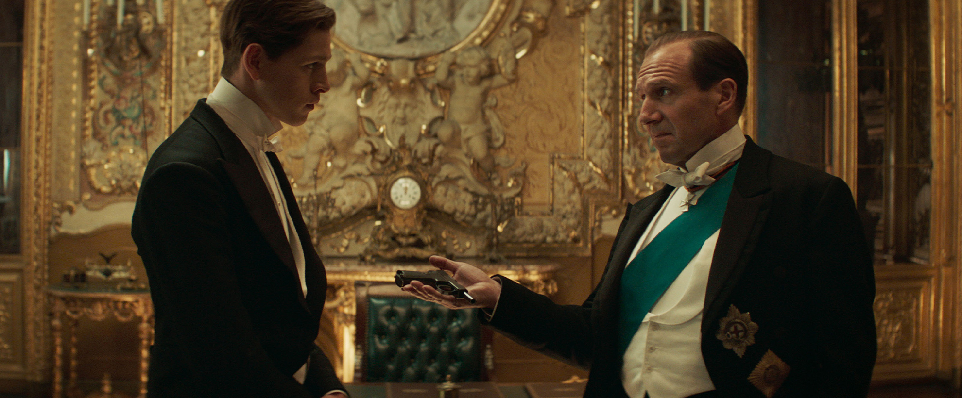 Ralph Finnes hands his son a pistol, in an opulent setting, dressed in formal best
