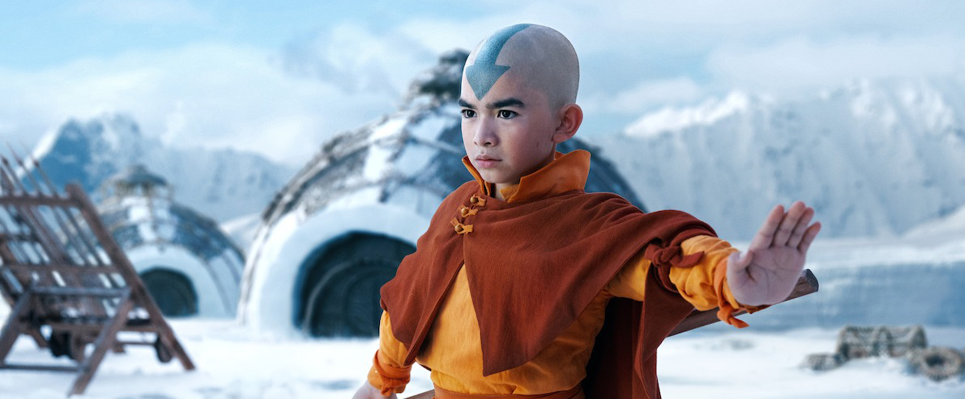 A young boy wearing orange robes, with a blue arrow painted on his shaved head, standing in a snowscape