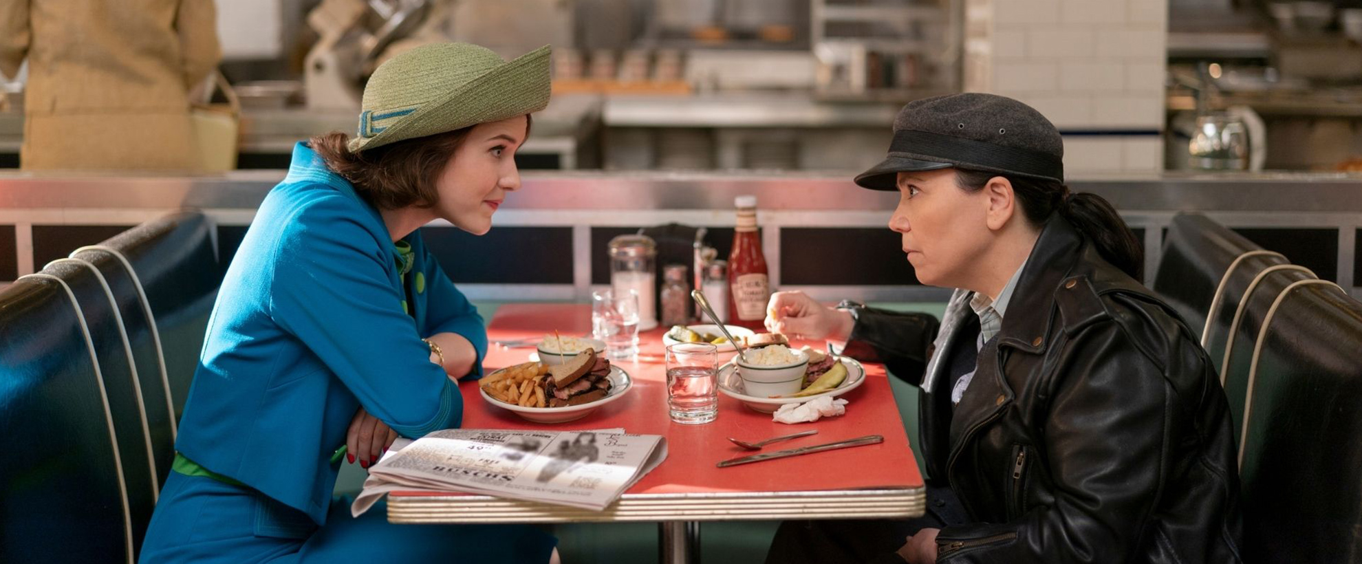 Mrs Maisel sits at a diner table, leaning over to speak with a person in a black leather jacket