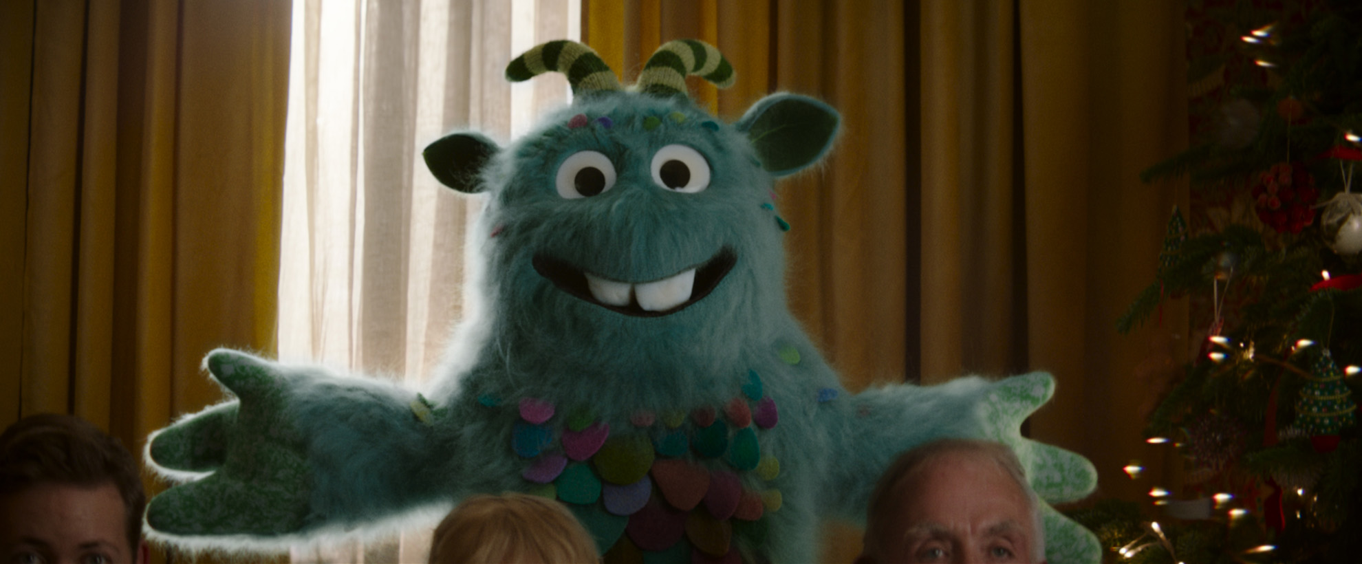 A blue fuzzy monster with stripey horns is standing in a living room, there are the foreheads of three people in the foreground.