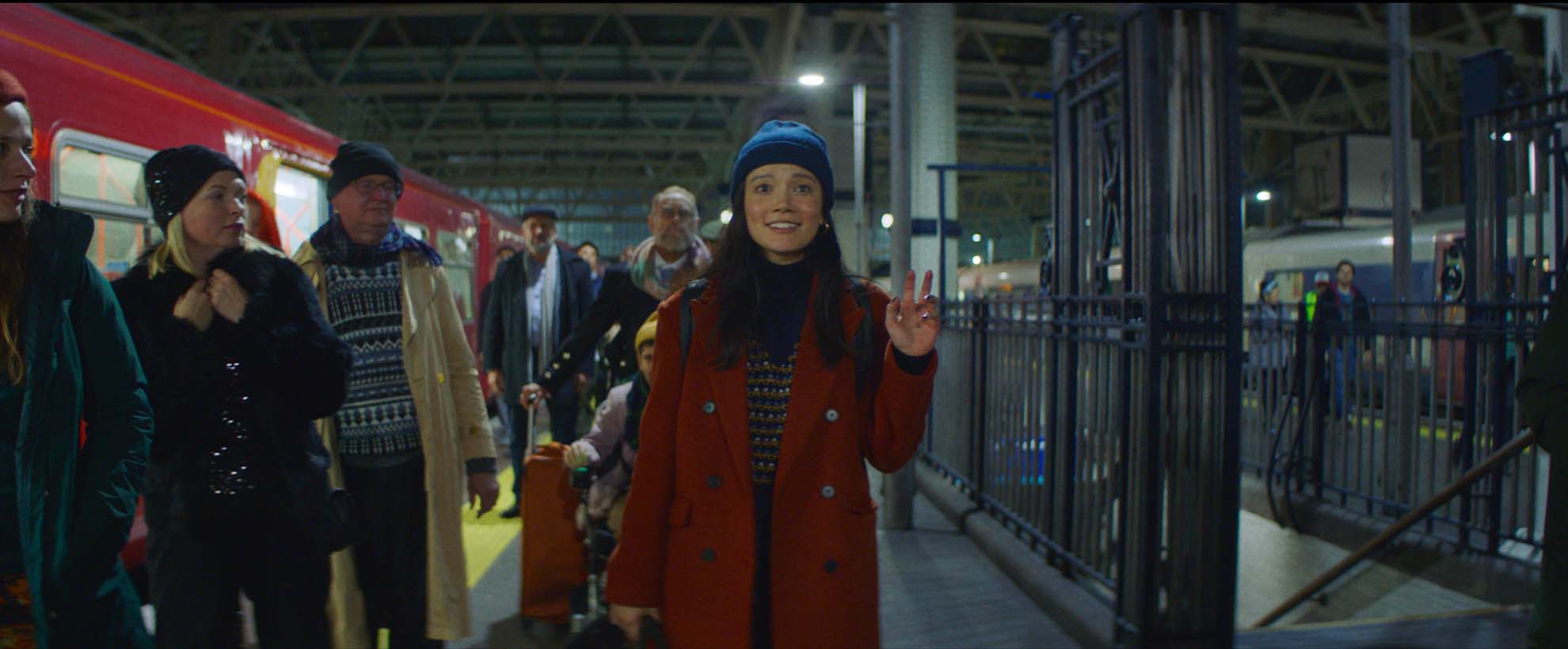 A woman in a red coat and blue winter beanie hat stands in a train station and waves
