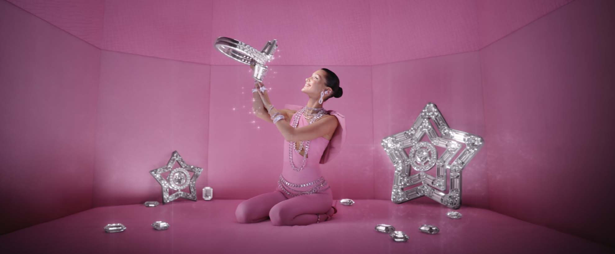 Bella Hadid sits in a pink room surrounded by large crystal stars holidng a large ring