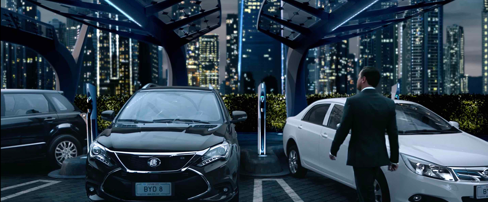 A man in a suit walks to a modern parking lot to his car. There is a city in the background. 