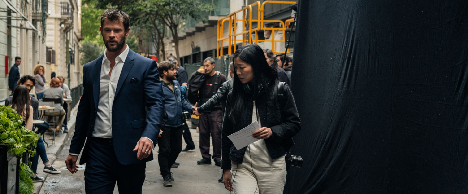 A man and a woman are walking down a street on a film set with many other people in the background.