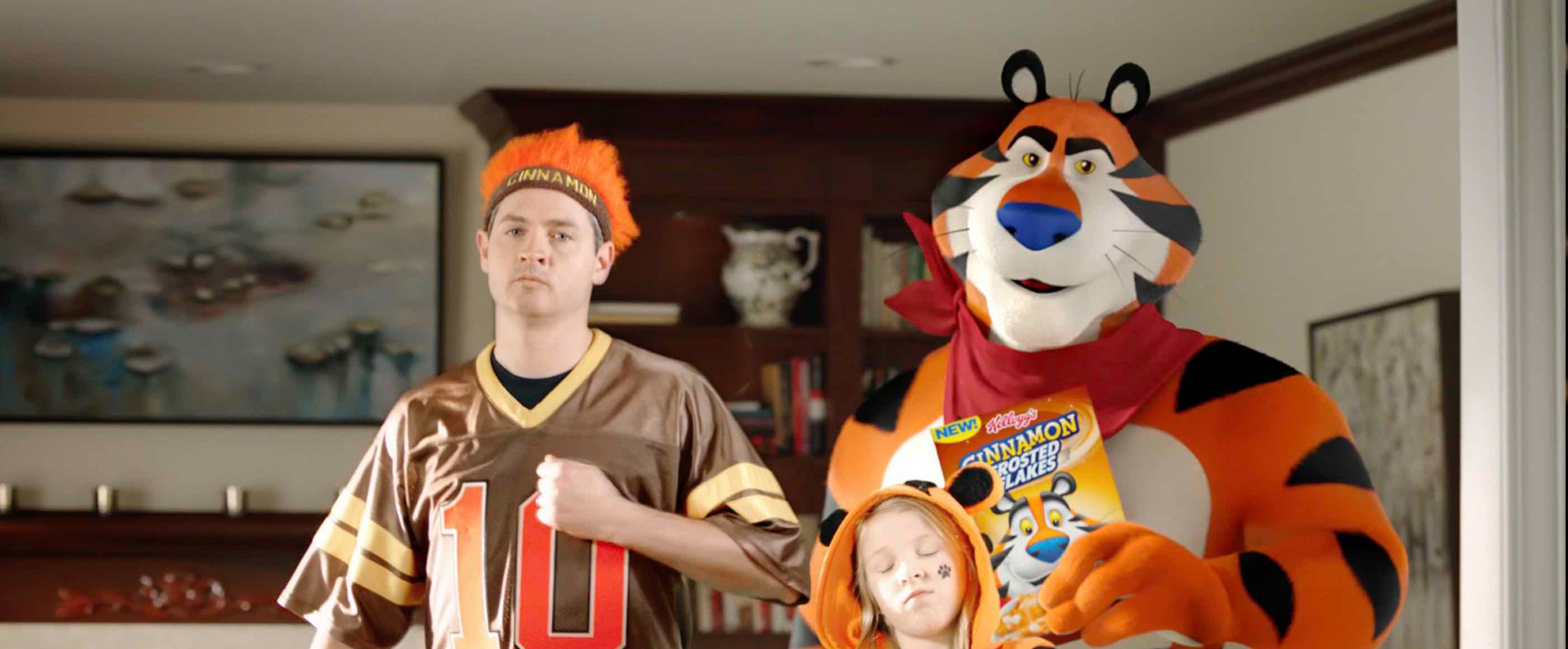 Tony the tiger holds a box of frosted flakes cereal and stands next to a young girl in a tiger hood and a man in a sports jersey
