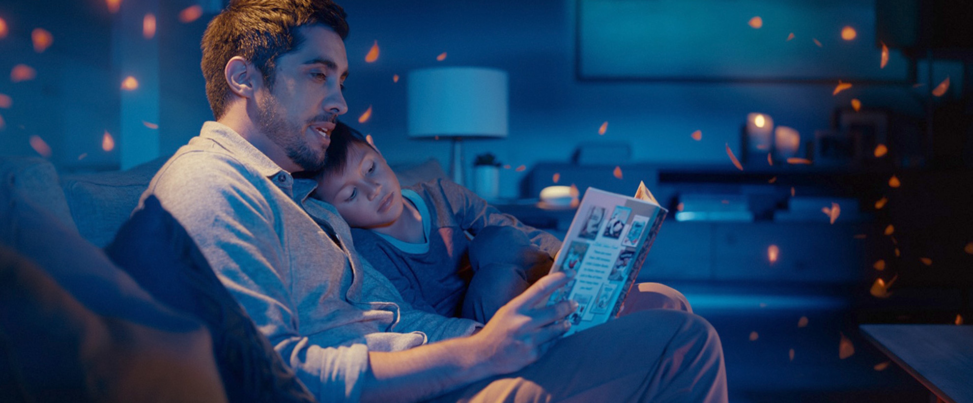 A man and his son sit on a sofa reading a book with a Google Home mini in the background.