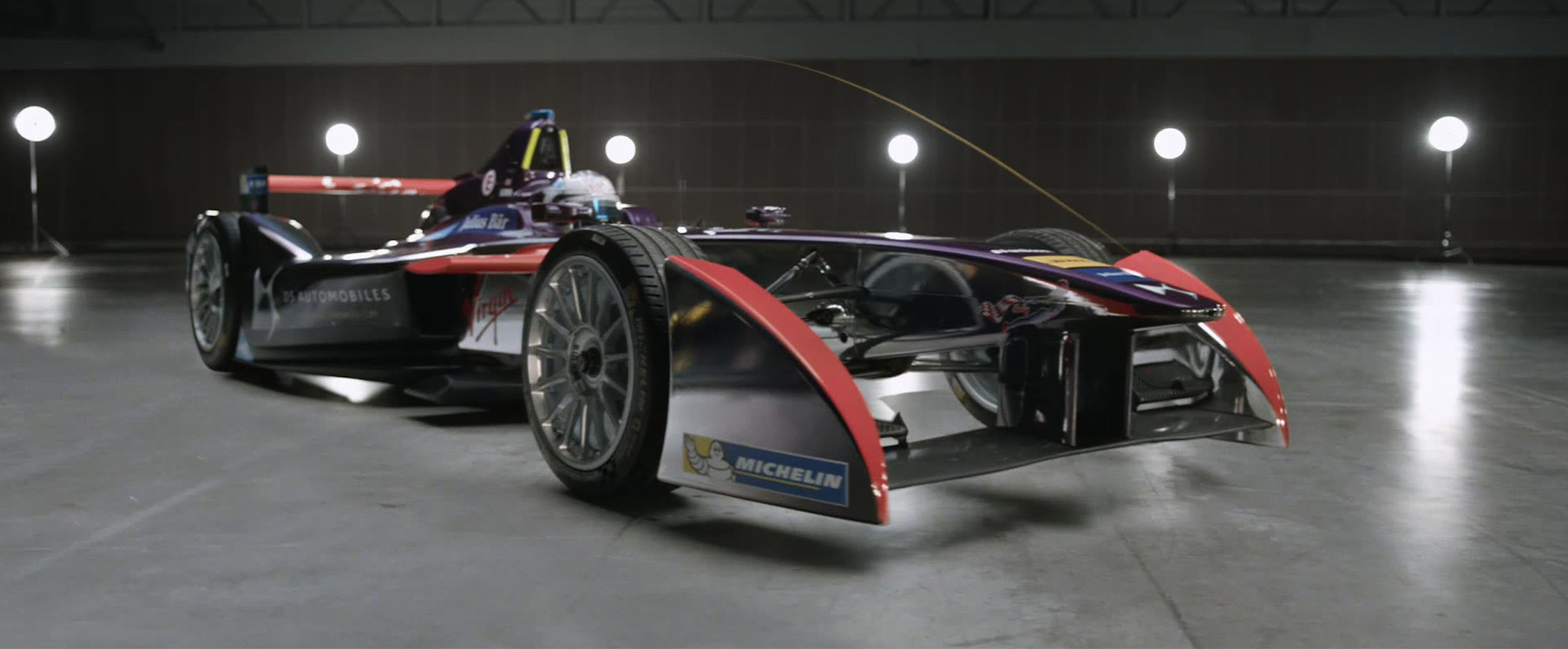 A formula E car with the front pointing towards the camera at a 45 degree angle, the car is in a large grey warehouse.
