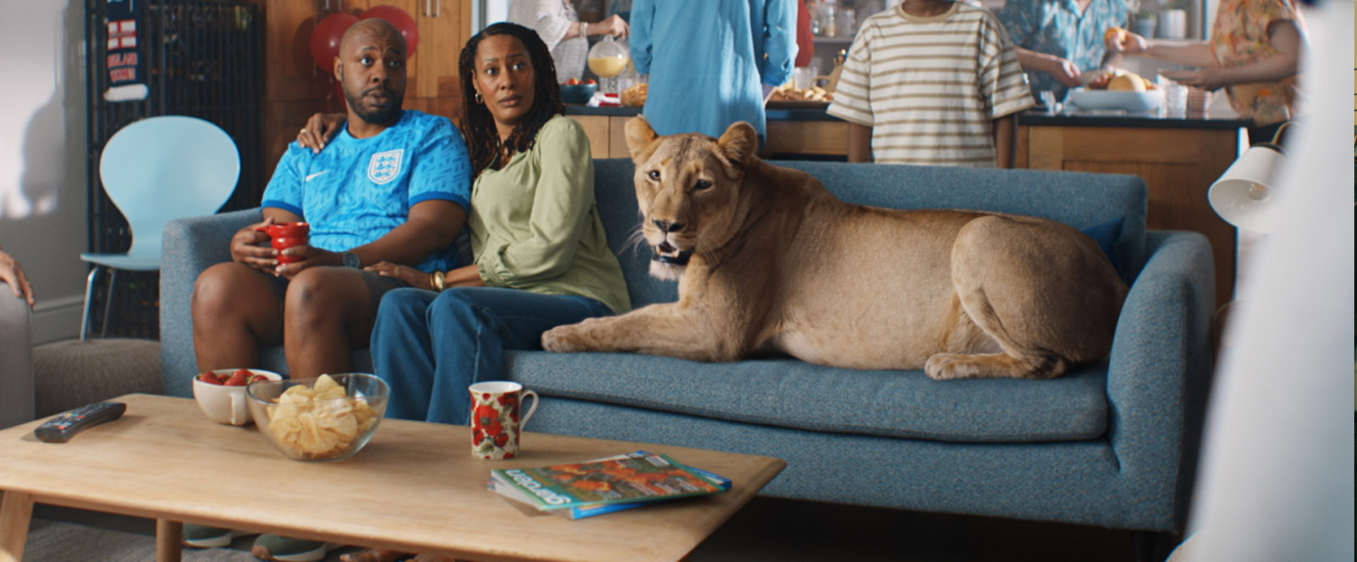 A lioness lays on a living room couch as a couple looks shocked sitting next to it