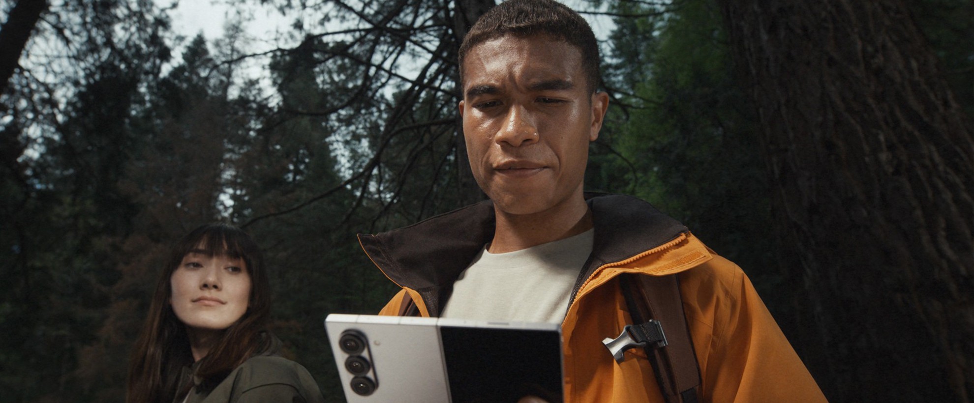 Two hikers, a man and a woman, in the woods. The man holds a Samsung Galaxy Z Fold6