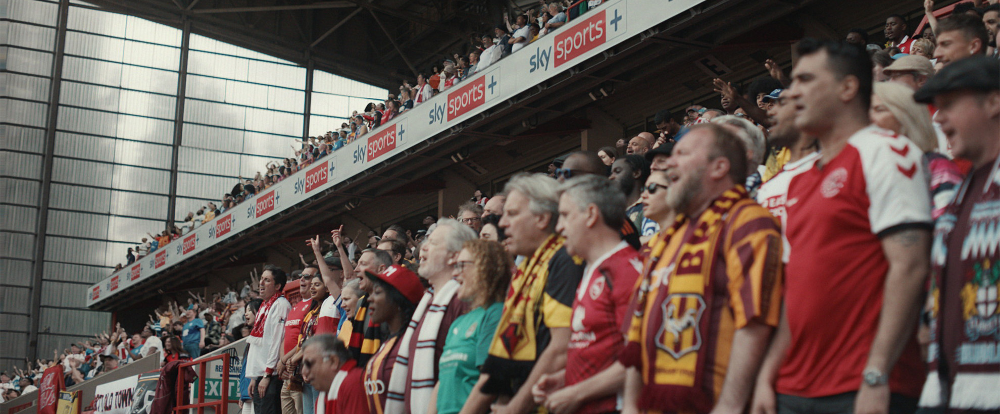 A crowd of football fans standing in the stands of the stadium with the Sky Sports banner above their heads. 