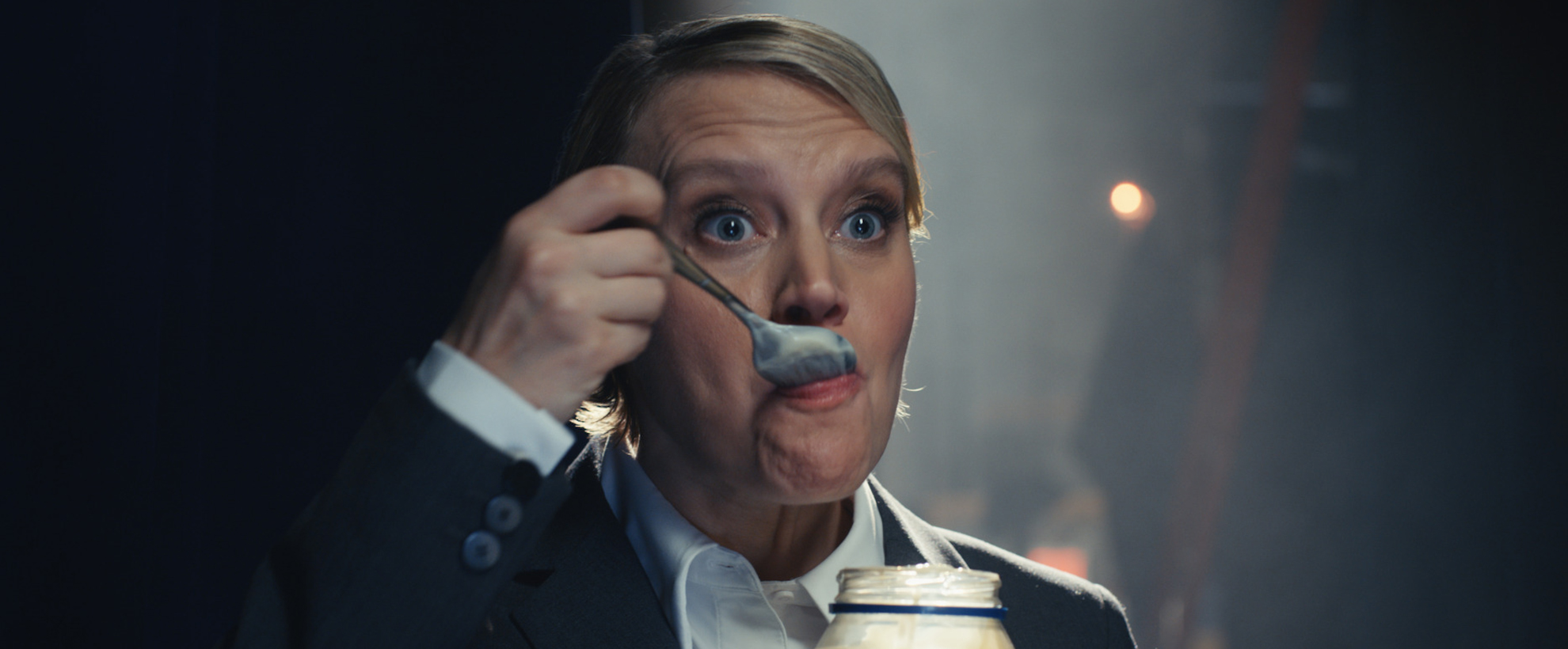 Actress Kate McKinnon on a stage in a suit eating mayonnaise from a jar