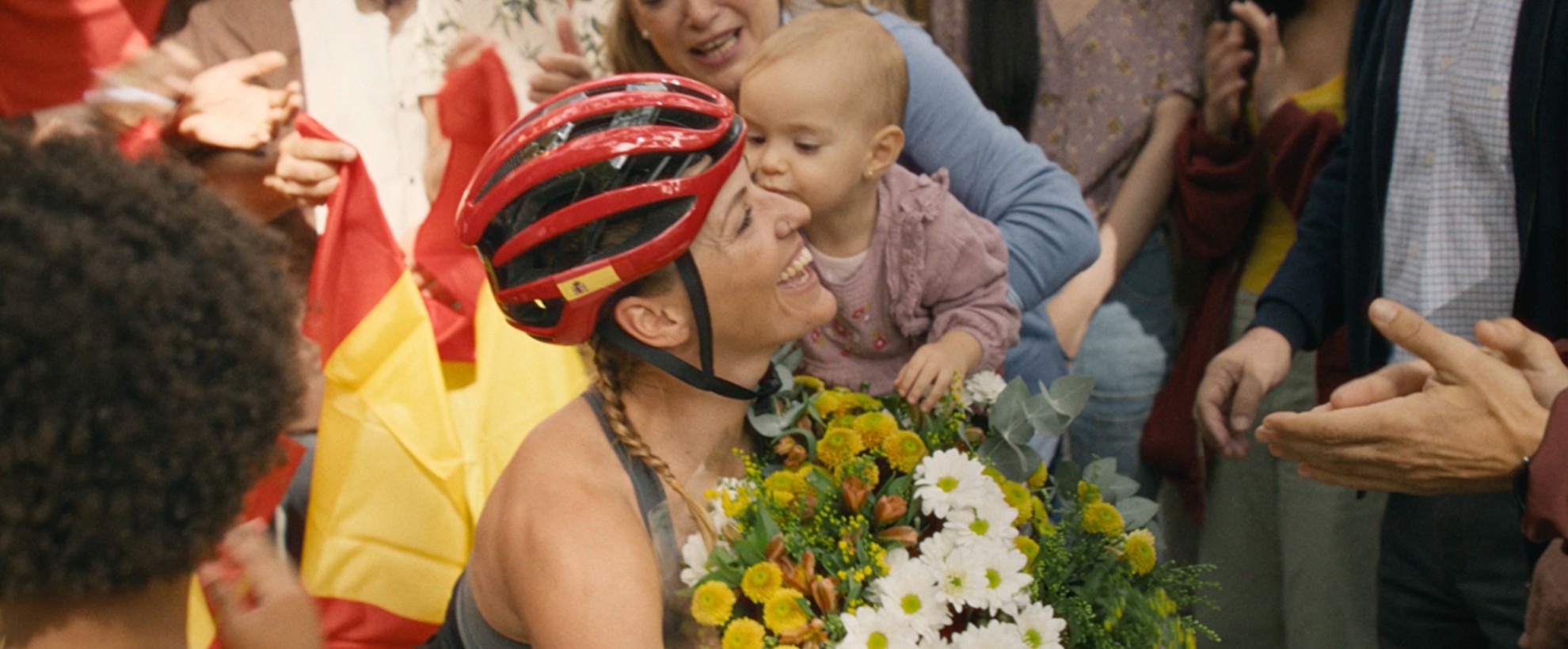 A Paralympian in her wheelchair bike holds a baby and a bouqet of flowers. A large group of loved ones and friends surround her after she completes her race. 