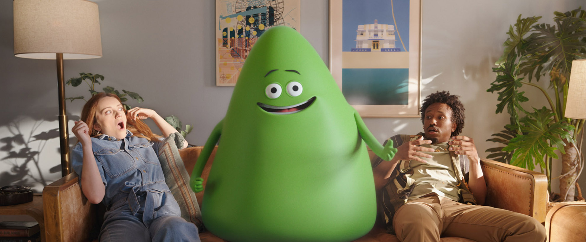 A large green Cricket mascot sits on a couch while two people look in awe