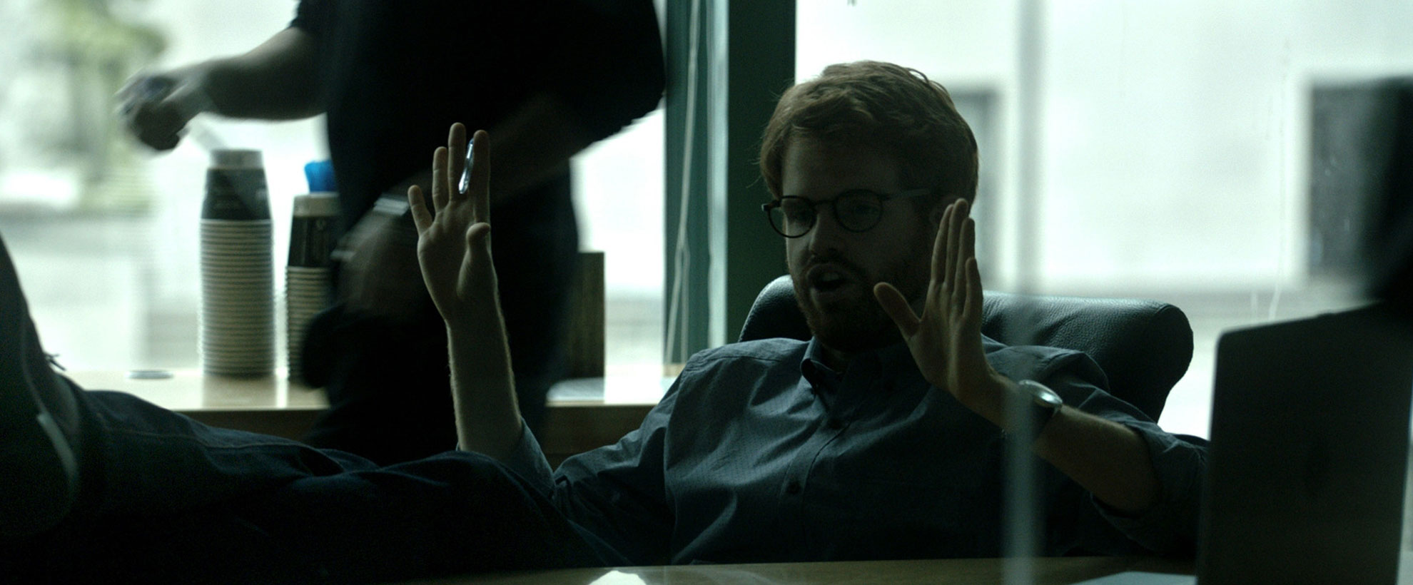 A man at a desk holds his hands up while on a conference call