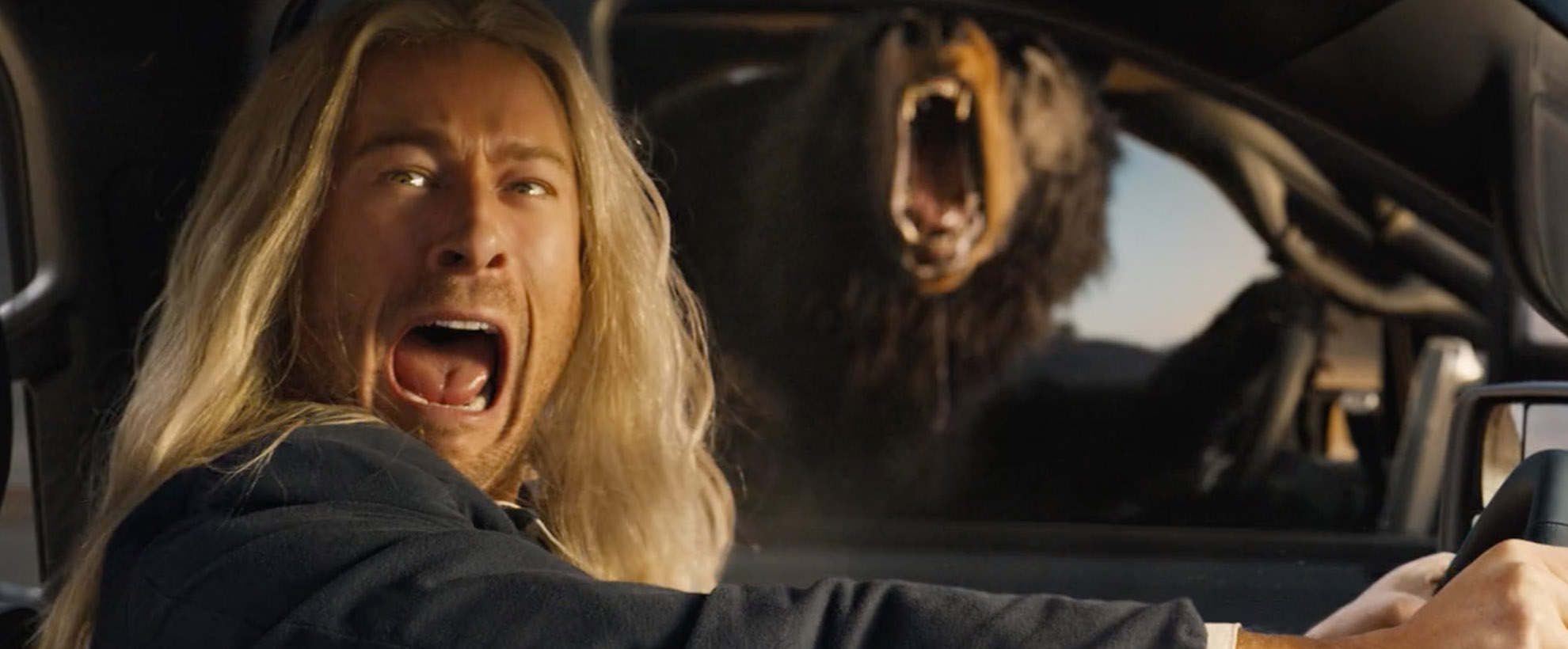 Glen Powell with long blonde flowing hair drives a truck and screams as he sees a (CG animated) bear in the vehicle next to him