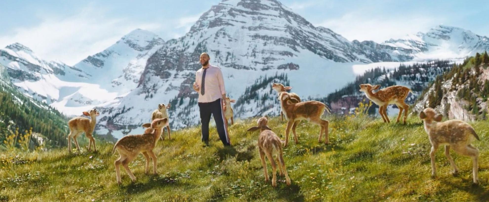Man surrounded by CGI deer standing in front of a mountain range 