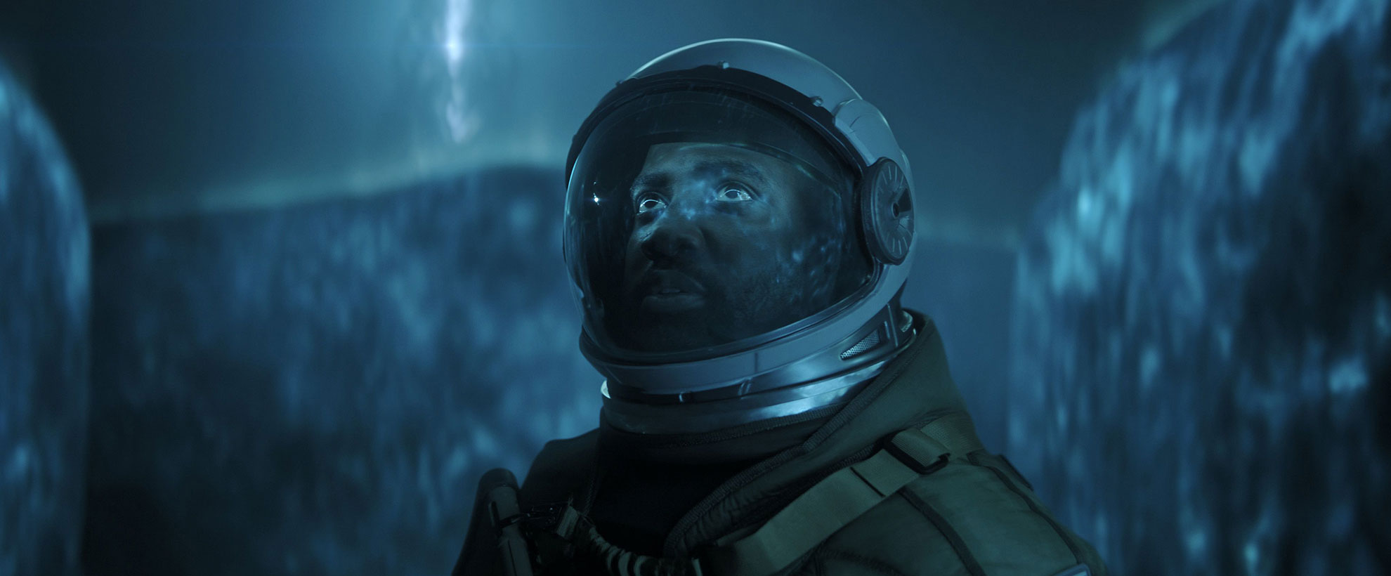 A person in a spacesuit stands in front of a neon blue extraterrestrial-like CGI gaseous wall with an alien entity's silhouette looming  