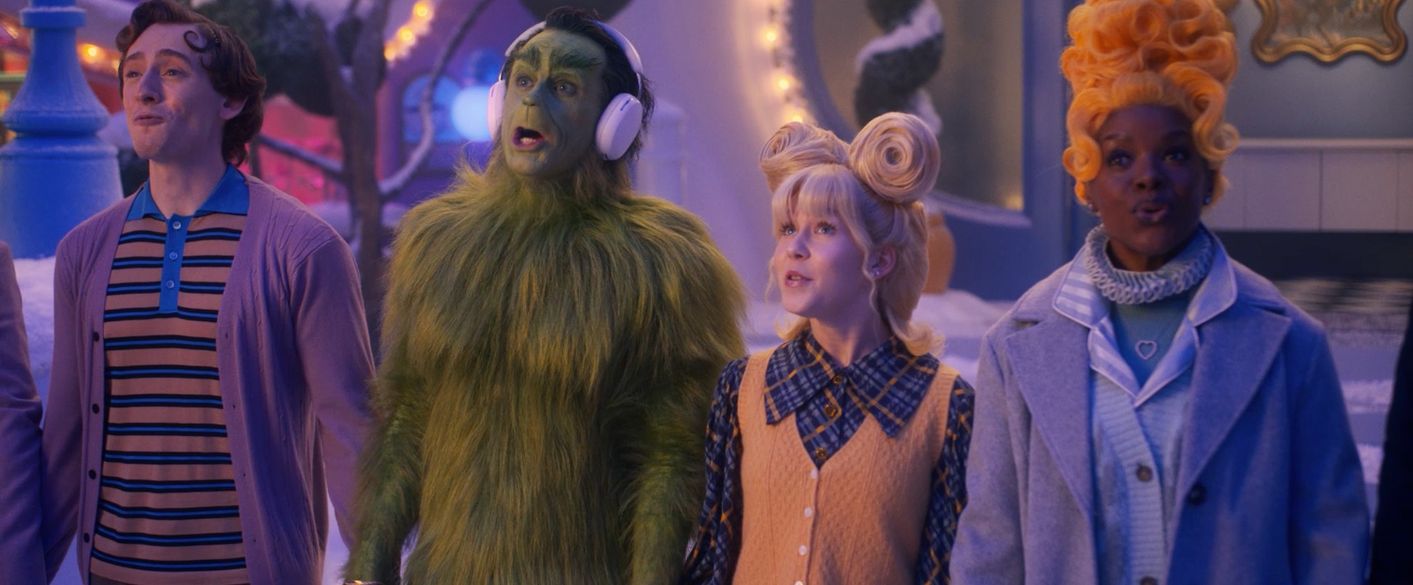 Four whimsical, brightly dressed characters—one being the Grinch with headphones—stand close together in a snowy Whoville, singing cheerfully.