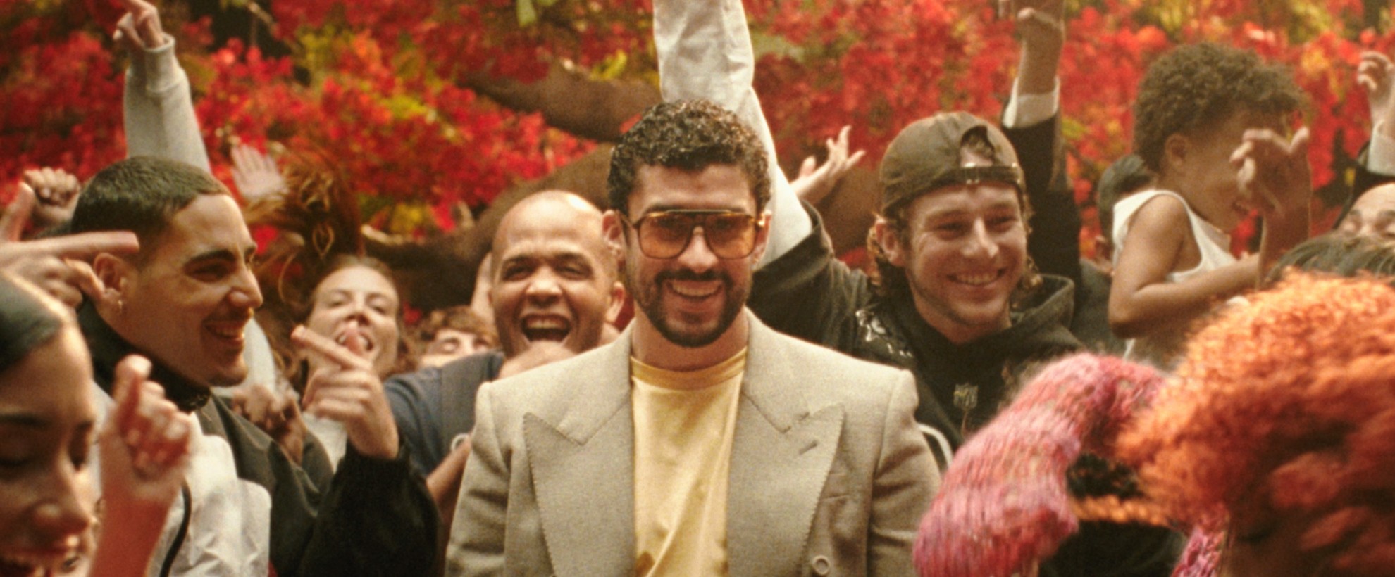 Bad Bunny in sunglasses and a blazer walks through a cheering crowd, arms raised, surrounded by red colored trees behind
