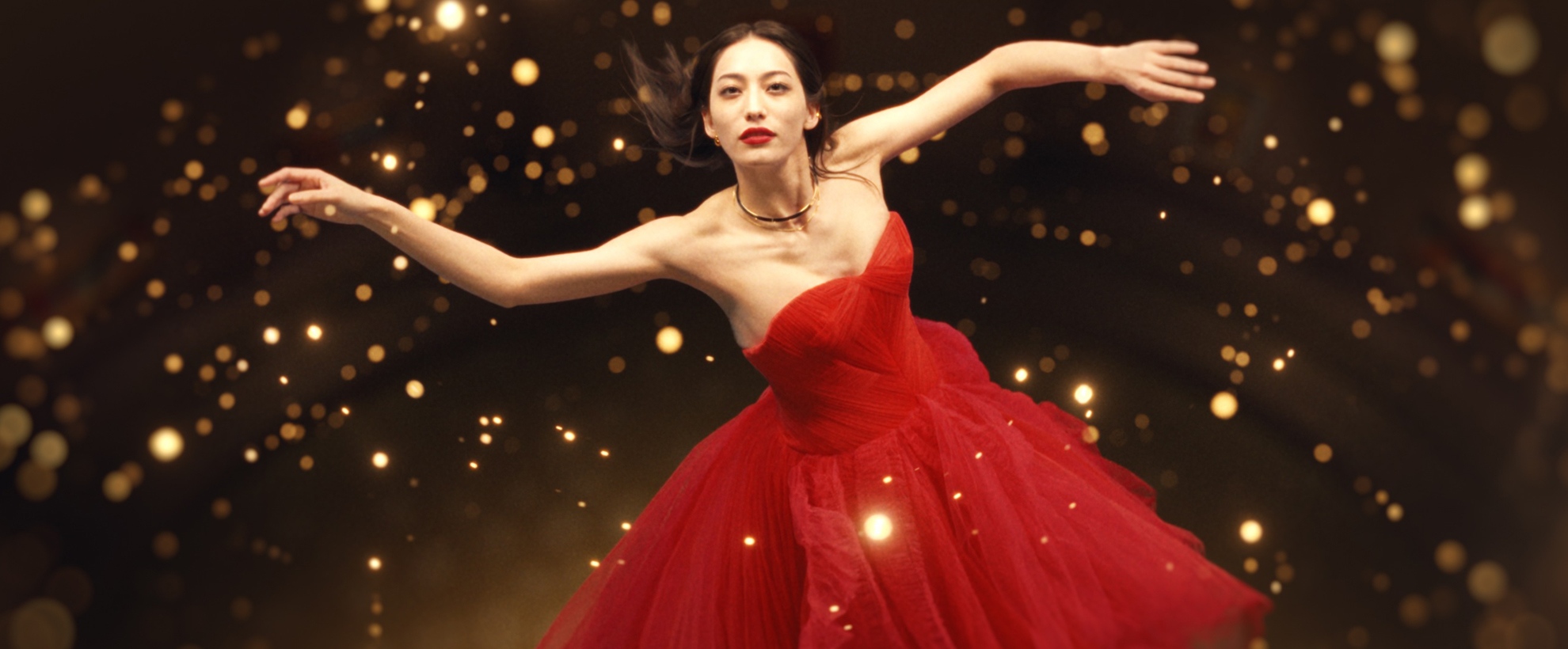 Woman in flowing red dress posing dramatically amid glowing golden lights against a dark background.