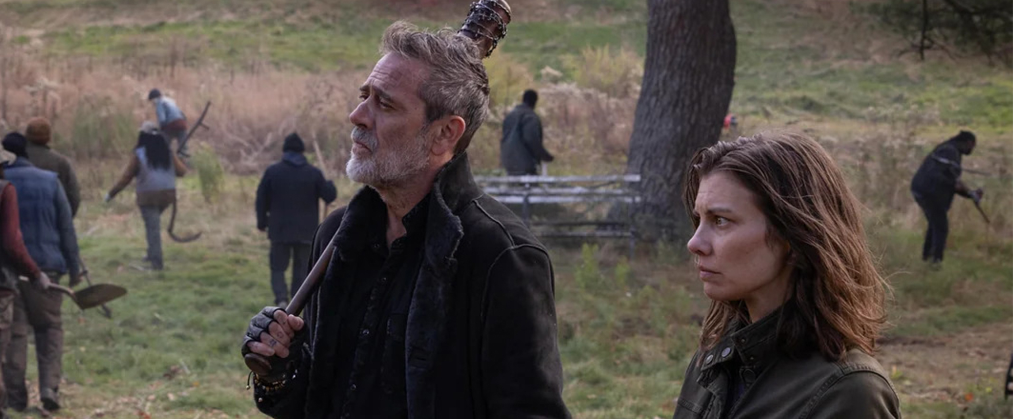 Negan and Maggie Greene from The Walking Dead: Dead City stand alert in a grassy field while others work in the background, creating a tense, post-apocalyptic atmosphere.