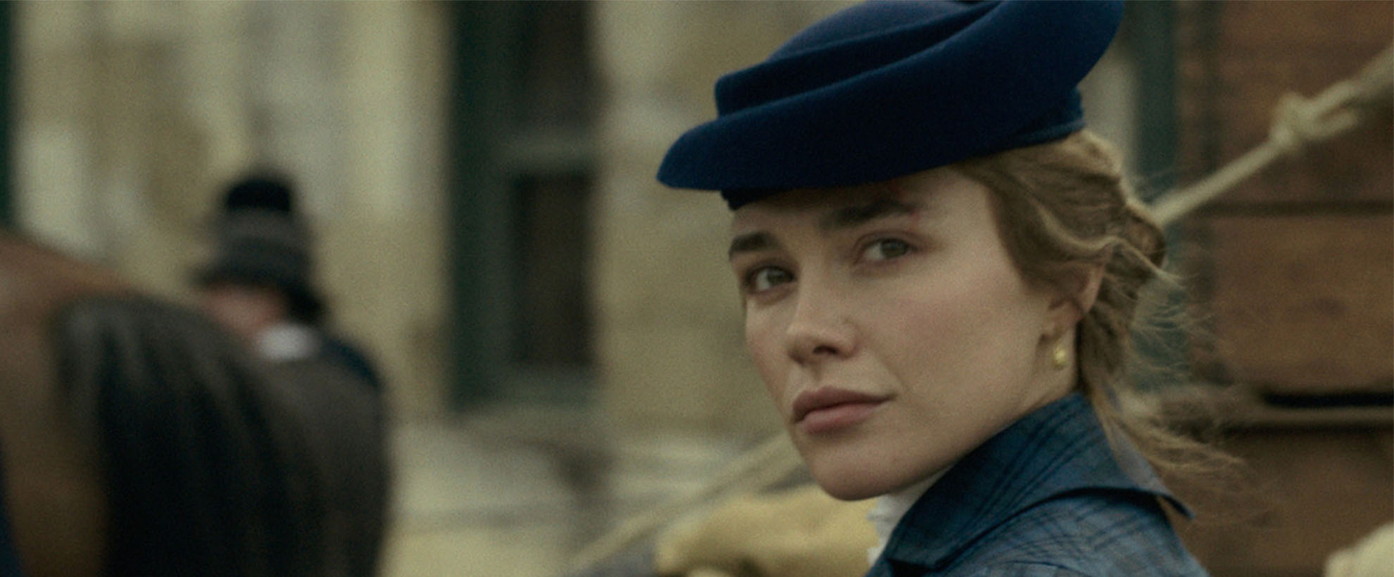 Florence Pugh as Cathy Ames in East of Eden, wearing period clothing and blue hat glances over her shoulder on a historic street, with background figures softly blurred.