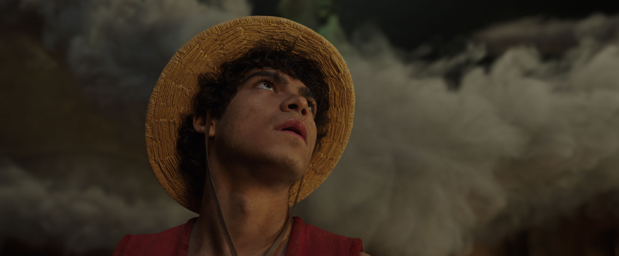 Luffy wearing a straw hat looks upward as thick smoke billows behind them in a dimly lit setting.