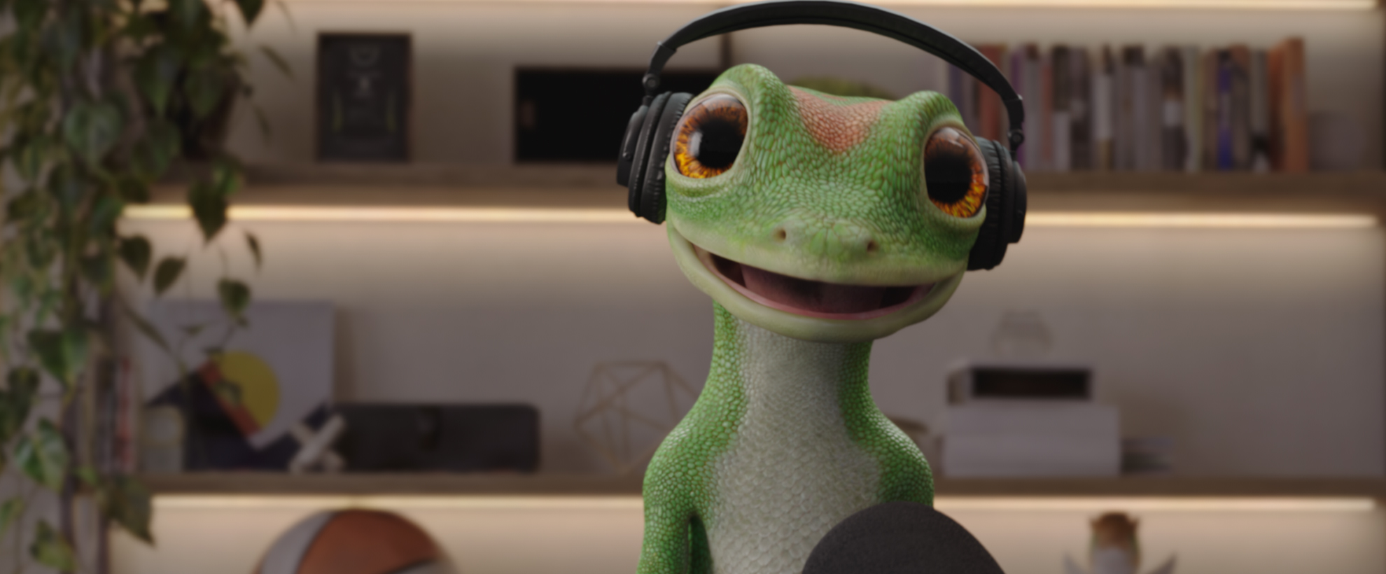 The CG animated GEICO Gecko wearing headphones speaks into a podcast microphone in a warmly lit studio with shelves, books, plants, and decorative items.