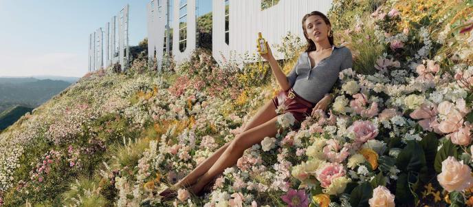 Miley Cyrus sits on a bed of flowers next to the Hollywood sign holding a bottle of Gucci fragrance