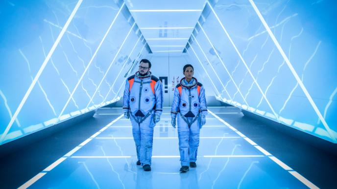 Two astronauts walk down a well lit walkway/tunnel