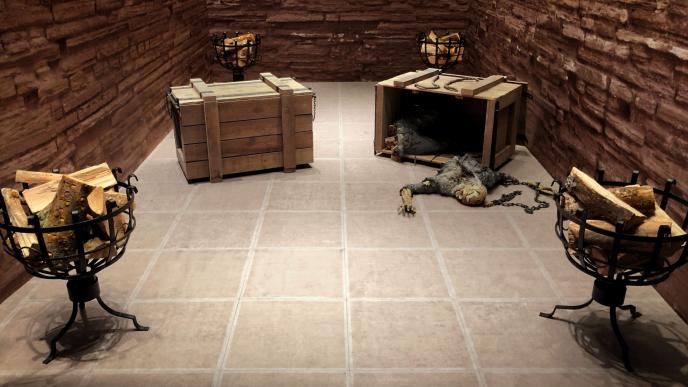 a brick room that has four iron log holders full of wood in each corner of the room. there are two boxes in the centre. one of them is closed. the other is open and has a decaying body in chains that has snapped in half