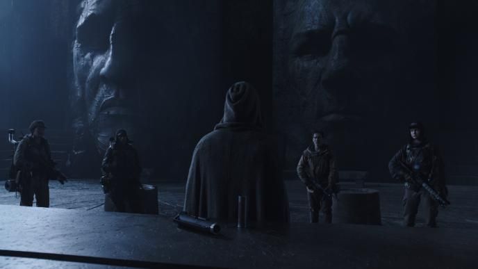back view of a person wearing a hooded cape standing in front of four armed soldiers facing them in a cave that has large engraved faces in it