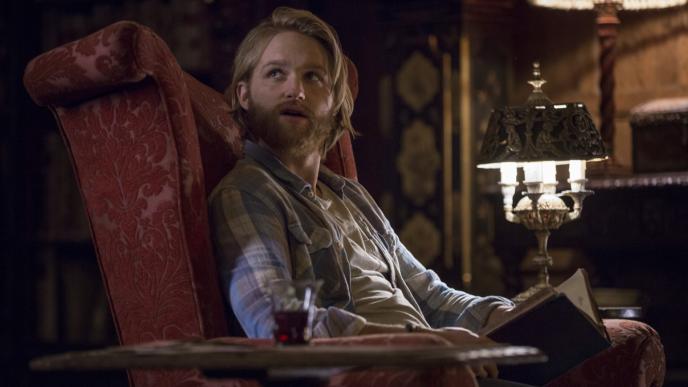 actor wyatt russell as cooper sitting in a detailed chair looking up to the left with an open book in his hand. there is a lamp shade and glass of red wine on either side of the chair