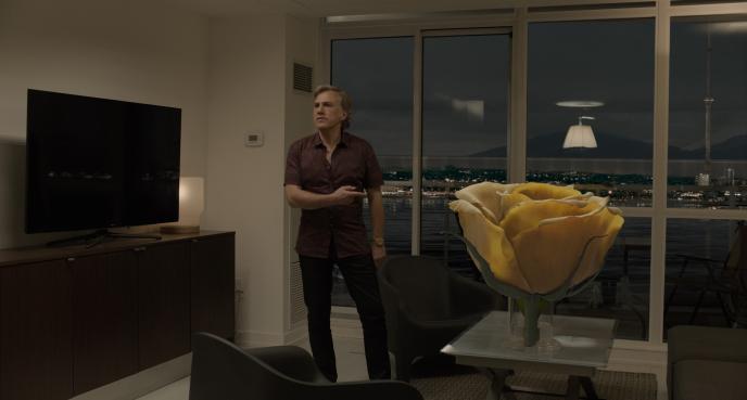 actor christopher waltz as dusan mirkovic standing in a living room pointing as a vase that has a giant yellow rose bud in it