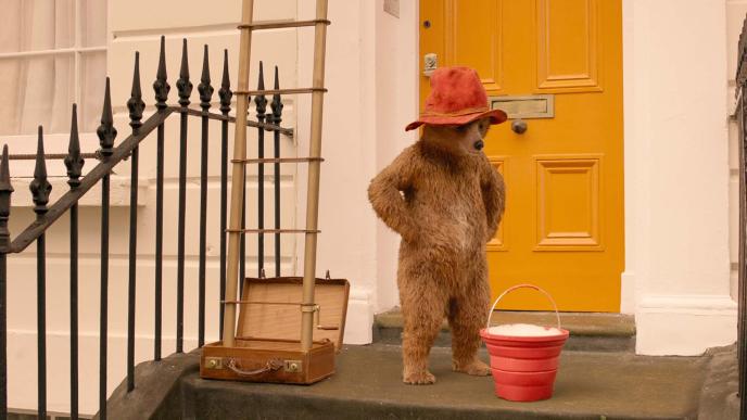 paddington bear wearing a red hat standing with his arms on his hips outside of a yellow door porch. he is looking down at a bucket full of soapy water. there is a ladder coming out of a suitcase on the left.