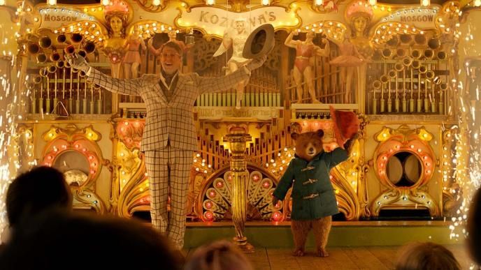 actor hugh grant as phoenix buchanan and paddington bear smiling while holding their arms and hats out in front of a fun fair