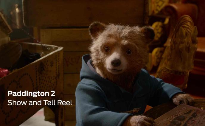 paddington bear wearing his blue button up coat sitting with his paw on the table. in the bottom left corner there is text that reads 'paddington 2 show and tell reel'