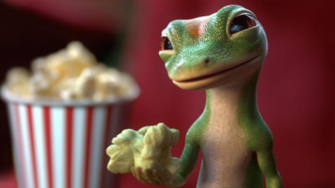 geico gecko holding up a piece of popcorn. there is a full popcorn box in the background