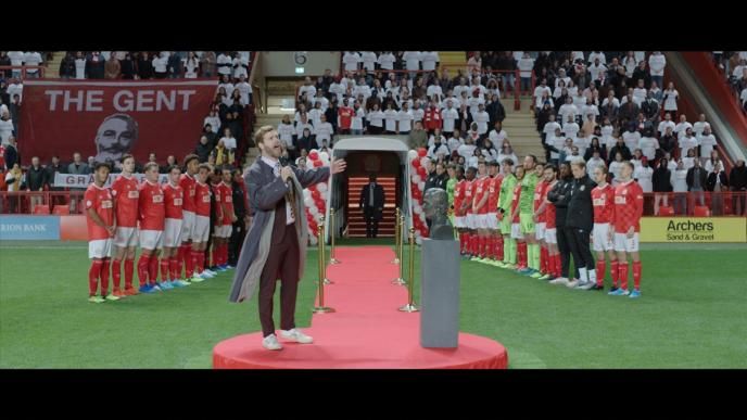 a commentator speaking into a microphone gesturing the two football teams standing in a 'v' formation on a football pitch as viewers stand on the bleachers