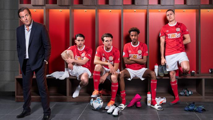 actor will barnet standing up in a locker room. there are four football players, three are sitting down and one is standing up against the lockers