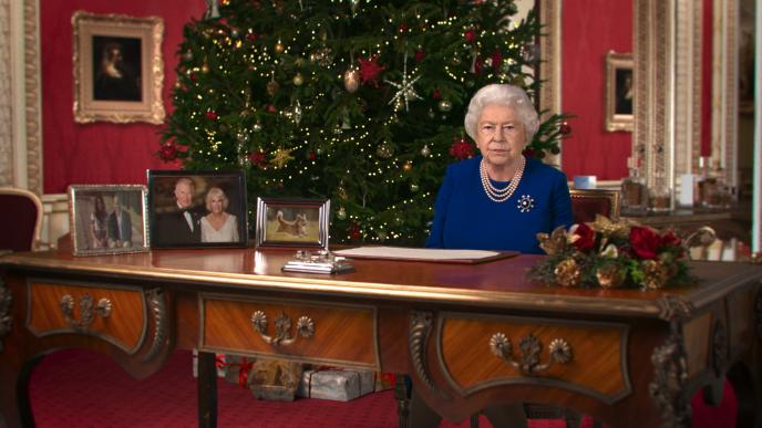 deepfake of queen elizabeth the second sitting behind a table. there is a large christmas tree behind her and the room is decorated festively