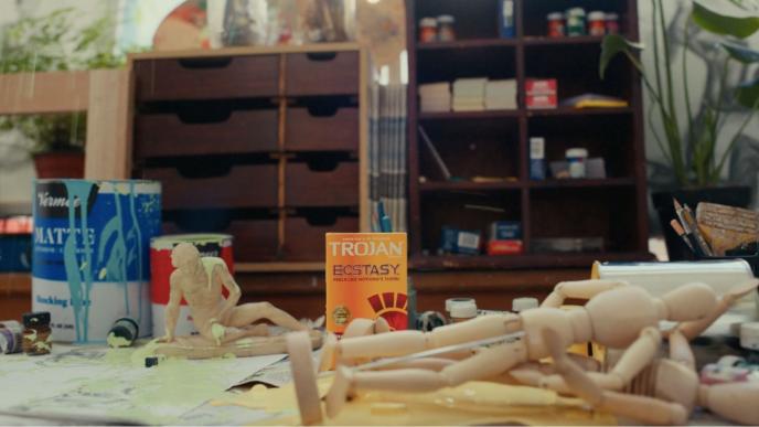 an artist table covered in paint and wooden mannequins. there is a box of trojan condoms in the centre of the table