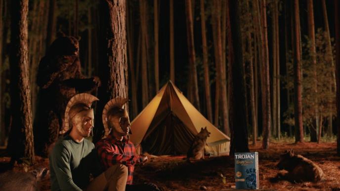 two people wearing roman helmets camping in the forest during the night. there is a bear approaching them from the side. a box of trojan condoms are on the right