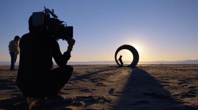 back perspective of a behind the scenes shot of a cameraman holding a camera as a director is directing an actor