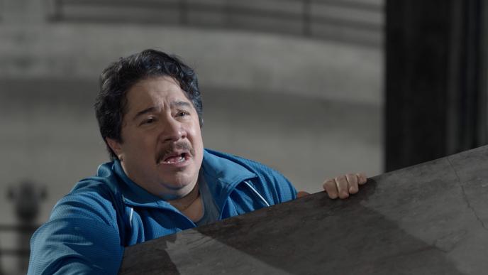 A mustached man dangles from the edge of a parking garage